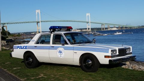 1991 Ford LTD Crown Victoria - Photograph of a white police sedan with blue striping parked at a harbor with a suspension bridge behind.