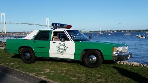 1991 Ford LTD Crown Victoria - Photograph of a green-and-white sheriff sedan with a star emblem parked at a waterfront with a suspension bridge.