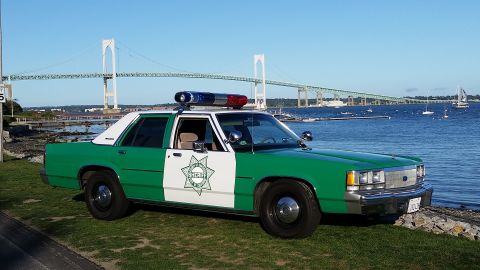 1991 Ford LTD Crown Victoria - Photo of a green-and-white sheriff Ford Crown Victoria police car parked along a bay with a suspension bridge behind it.