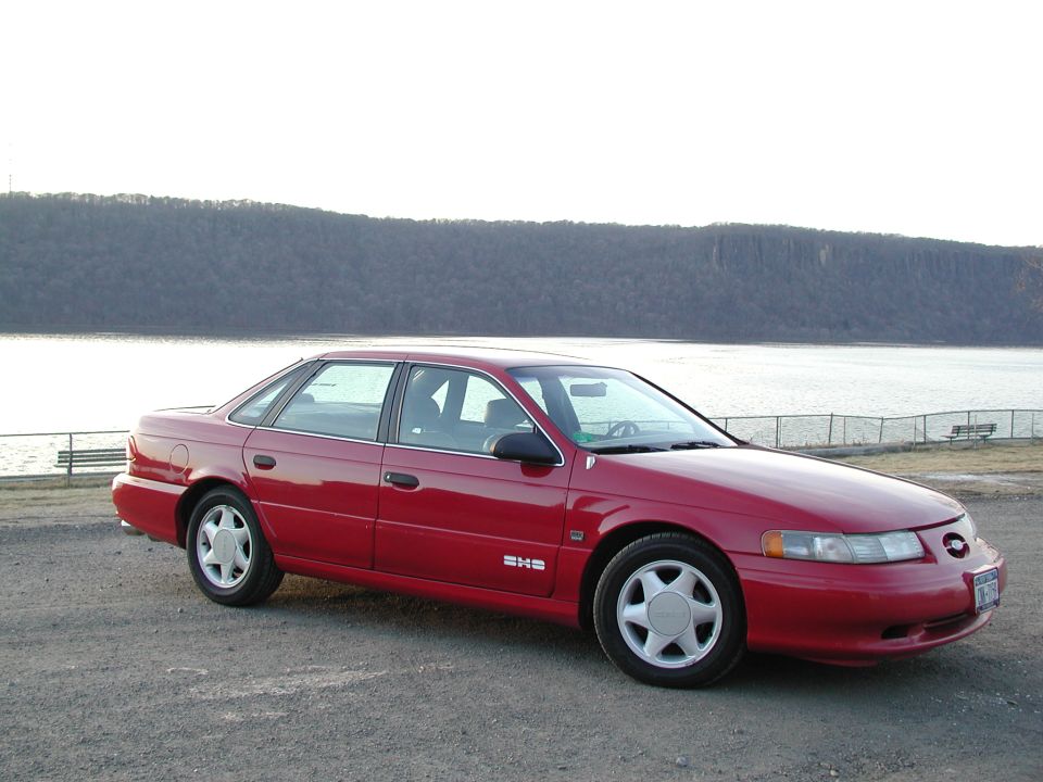 Red Ford Taurus SHO sedan parked on gravel by a lake with a wooded bluff behind