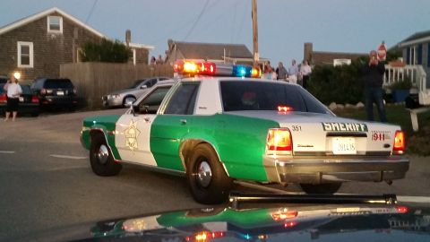 1991 Ford LTD Crown Victoria - Photo of the rear of a green-and-white sheriff Ford Crown Victoria with emergency lights on, spectators watching at dusk.