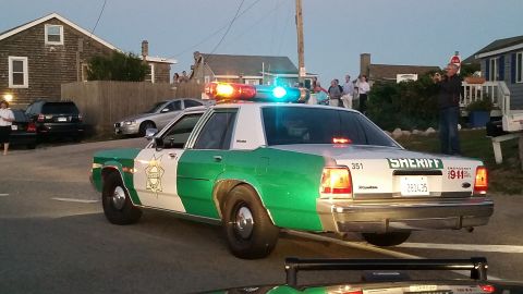 1991 Ford LTD Crown Victoria - Photo of a green-and-white sheriff Crown Victoria with flashing lights and a crowd of onlookers on a coastal street at dusk.