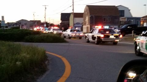 1991 Ford LTD Crown Victoria - Photo of a line of police cruisers with flashing lights on a curving coastal road at dusk.