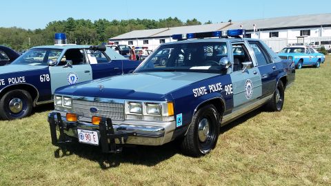 1991 Ford LTD Crown Victoria - A two-tone blue and grey 1991 Ford Crown Victoria police car is parked on a grassy field with a building and trees in the background. The c…