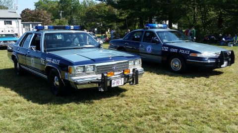 1991 Ford LTD Crown Victoria - Two Ford Crown Victoria police cars are parked on a grassy field on a sunny day. The car in the foreground is dark blue and light blue with…