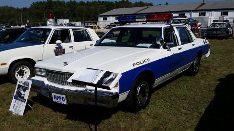 1991 Ford LTD Crown Victoria - A white and blue police car with a light bar on top is parked on a grassy field. A sign in front of the car displays information about a po…