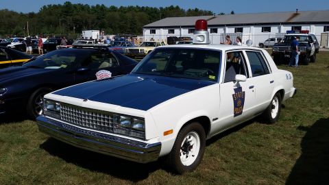 1991 Ford LTD Crown Victoria - A white and blue 1980s era police sedan with a red light bar on top is parked on grass at an outdoor event. The car has "STATE POLICE" deca…