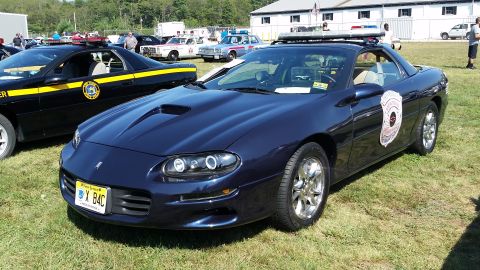 1991 Ford LTD Crown Victoria - A dark blue Chevrolet Camaro police car is parked on a grassy field with other vehicles in the background. The car has a light bar on the r…