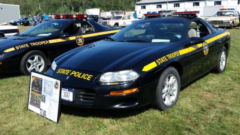 1991 Ford LTD Crown Victoria - Two black Chevrolet Camaros with yellow "STATE TROOPER" and "STATE POLICE" decals are parked on a grassy area. A display board with informa…