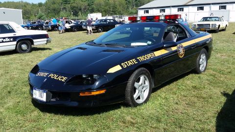 1991 Ford LTD Crown Victoria - A black 1990s Chevrolet Camaro Z28 convertible police car with yellow "STATE POLICE" and "STATE TROOPER" decals is parked on a grassy field…