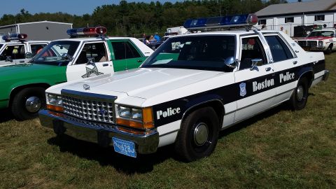 1991 Ford LTD Crown Victoria - A white-and-dark-blue 1980s Ford LTD Crown Victoria police cruiser lettered 'Boston Police,' parked on grass at a car show; front three-qua…