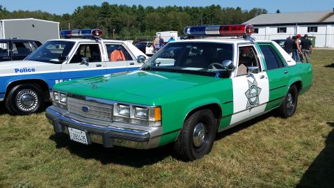 1991 Ford LTD Crown Victoria - A green and white Ford Crown Victoria police car with a light bar is parked on grass in front of a building. The car has "SHERIFF" written…