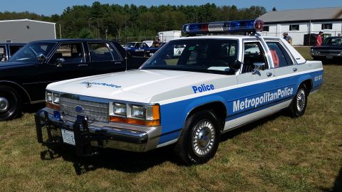 1991 Ford LTD Crown Victoria - A white and blue 1991 Ford Crown Victoria police car is parked on a grassy field with trees in the background. The car has "Metropolitan Po…