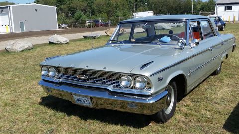 1991 Ford LTD Crown Victoria - A silver 1960s era Ford sedan is parked on a grassy area with a large industrial building in the background. The car has a chrome grille, d…