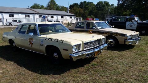 1991 Ford LTD Crown Victoria - Two vintage police cars, a cream-colored Plymouth Fury and a tan Ford LTD, are parked on a grassy field.