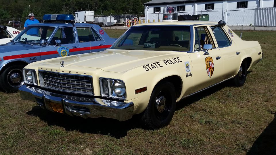 Cream-colored Plymouth Gran Fury police car with 'STATE POLICE' on the door, parked next to a blue and red police car