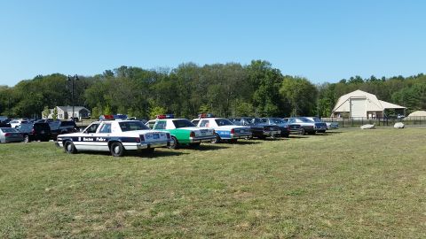 1991 Ford LTD Crown Victoria - Several police cars, including a 1991 Ford Crown Victoria, are parked in a grassy field on a sunny day.