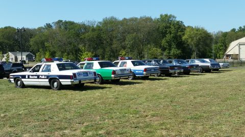 1991 Ford LTD Crown Victoria - A lineup of police vehicles and other cars parked on a grassy field under a clear blue sky.
