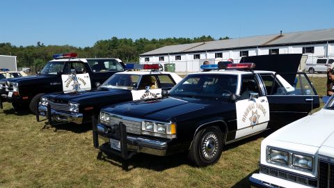 1991 Ford LTD Crown Victoria - A lineup of vintage highway patrol vehicles, including a black Dodge Ram and several sedans, are displayed outdoors.