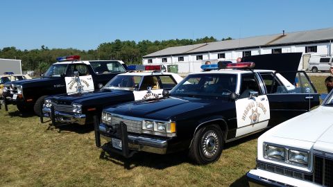 1991 Ford LTD Crown Victoria - Several police cars, including a black and white model, are displayed outdoors on a sunny day.