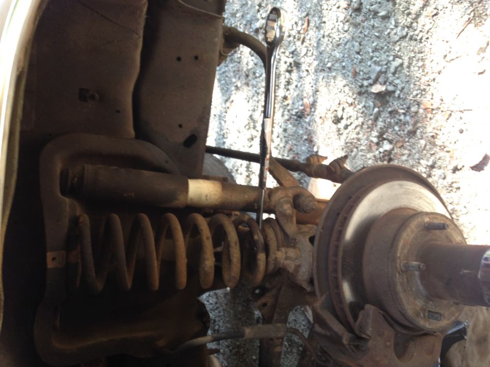close-up of a vehicle's front suspension with a coil spring, shock absorber, and brake rotor visible. A wrench is inserted into the suspension components