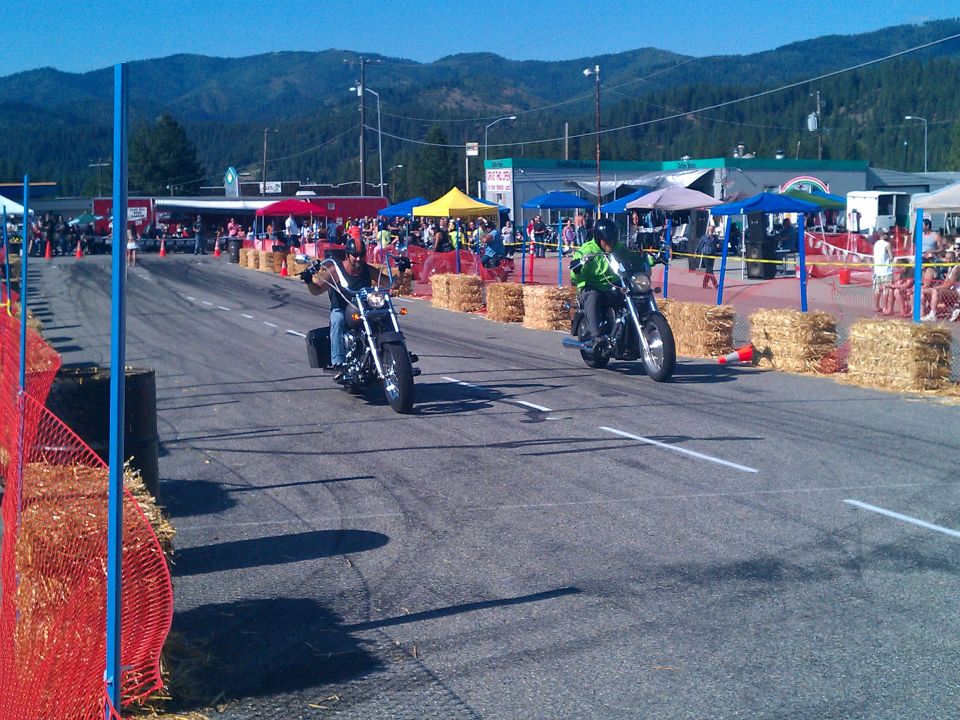 Two motorcycles racing on a street with spectators and hay bales