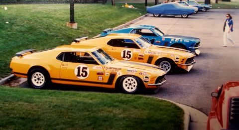 1973 Ford Mustang - Several vintage race cars are lined up on pavement next to a grassy area.
