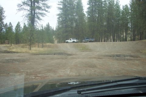 1995 Ford Bronco - Two pickup trucks drive on a dirt road surrounded by trees.