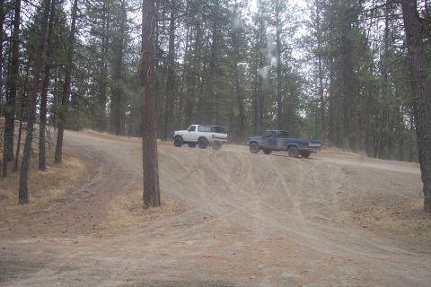 1995 Ford Bronco - Two off-road vehicles, a white 1995 Ford Bronco and a blue pickup truck, navigate a dusty trail through a wooded area.