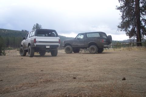 1995 Ford Bronco - Two Ford Broncos are parked on a dirt lot with trees and hills in the background.