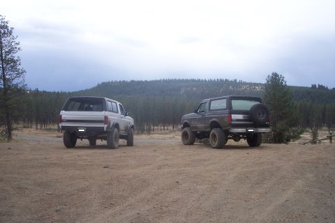 1995 Ford Bronco - Two Ford Broncos, a white one and a dark one, are parked in a dirt lot with a wooded hillside in the background.