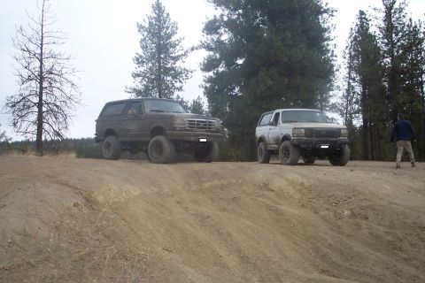 1995 Ford Bronco - Two Ford Broncos are parked on a dirt incline with trees in the background.