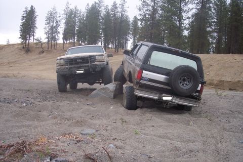 1995 Ford Bronco - Two Ford Broncos are shown off-roading on a dirt and rock path with trees in the background.