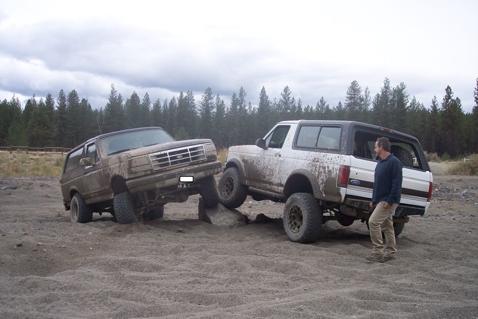 Muddy brown and white Ford Broncos are off-roading on a dirt path with trees in the background. A man stands near the white Bronco