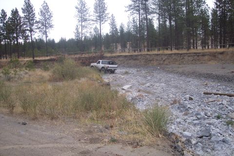 1995 Ford Bronco - A white SUV navigates a dry, rocky creek bed surrounded by trees and a wooden fence.