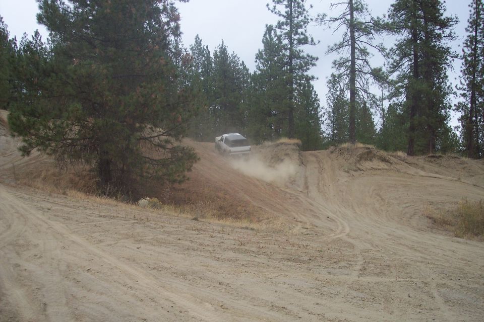 White SUV kicking up dust while driving up a dirt hill with pine trees in the background