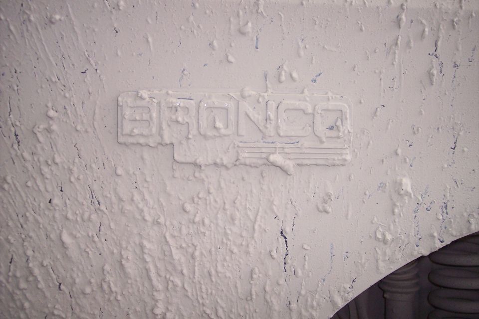 Embossed "BRONCO" text on a rough, painted surface. Part of a tire visible at bottom right