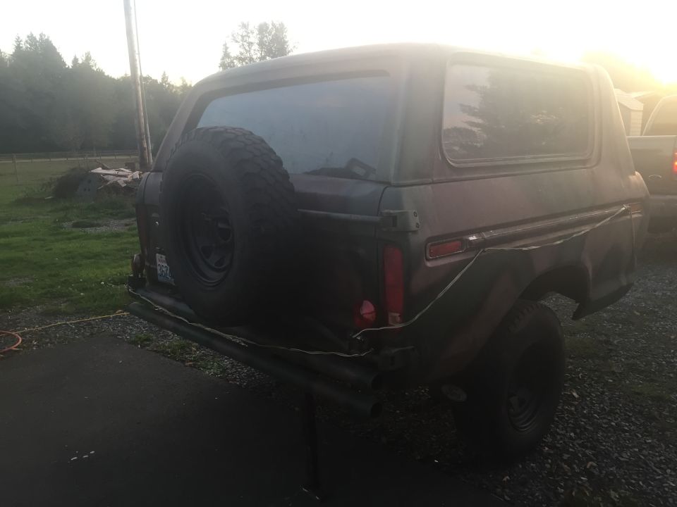 Rear view of a 1978 Ford Bronco with a spare tire mounted on the back. It's parked on a driveway with a grassy area and trees in the background