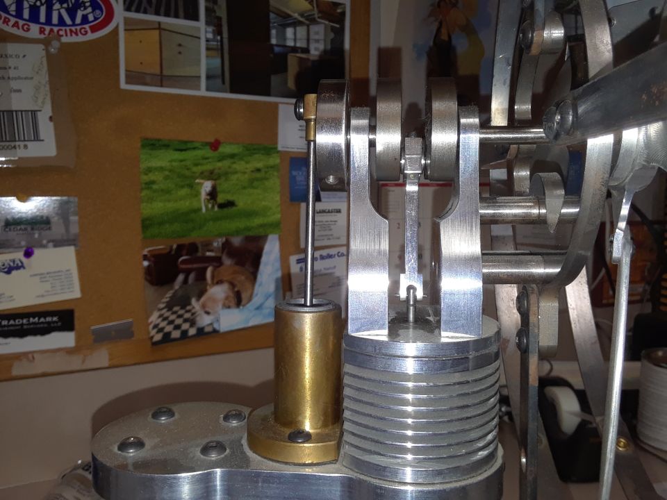 Close-up of a model engine's crankshaft and piston mechanism. Photos and labels are pinned to a corkboard in the background