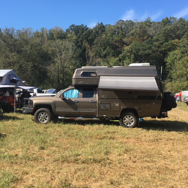 Brown truck with a camper shell parked in a grassy field with trees in the background