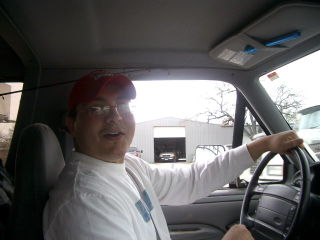 Man in red hat and glasses driving. Building with a truck inside is visible through the windshield