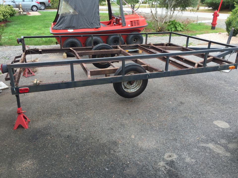 Utility trailer frame with wheels, supported by red jack stands. Red amphibious vehicle in background
