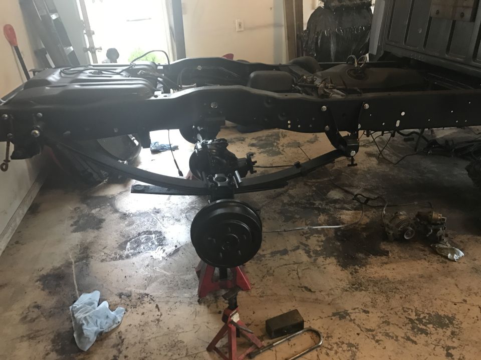 Truck frame with leaf springs and rear axle on jack stands. Black painted metal, some fluid stains on concrete floor