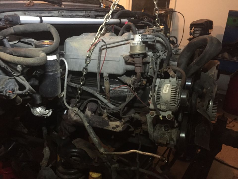 Engine assembly suspended by chains, showing alternator, hoses, and wiring. Some parts are dirty and worn