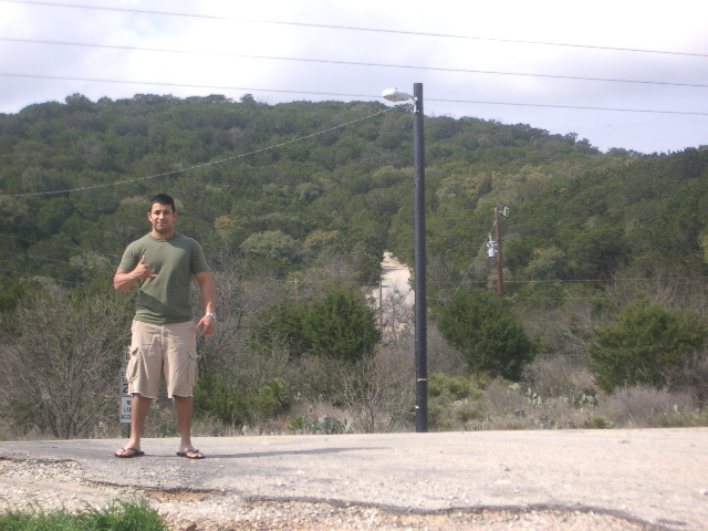 Man in green t-shirt and khaki shorts stands on a dirt road with a wooded hill behind him