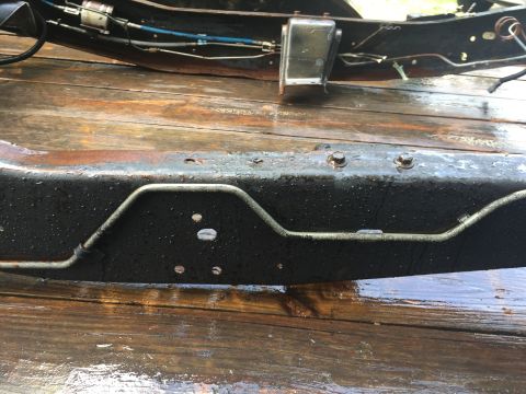 1995 Ford F-150 - A close-up view of a rusty, black truck frame with water droplets on it, resting on a wet wooden surface. Metal lines and bolts are visible…