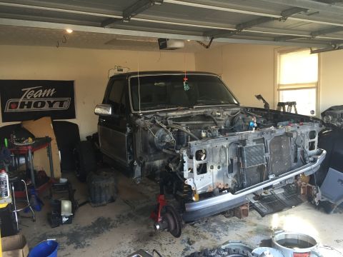 1995 Ford F-150 - A black 1990s Ford F-150 pickup truck is disassembled in a garage, with the front end removed and parts scattered around the floor. The tru…