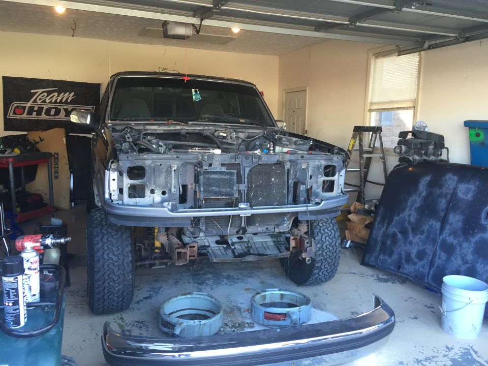 Black Ford F-150 pickup truck in a garage with hood removed, engine exposed, parts on floor.