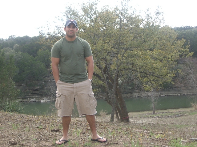 Man in green t-shirt and khaki shorts stands by a lake with trees in the background