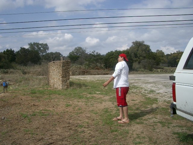 Guy in red shorts and white shirt stands by a white SUV. Stone structure in background